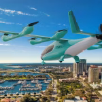 Electric VTOL aircraft flying over a coastal city with a marina and tall buildings below, blue sky overhead, cityscape in the background